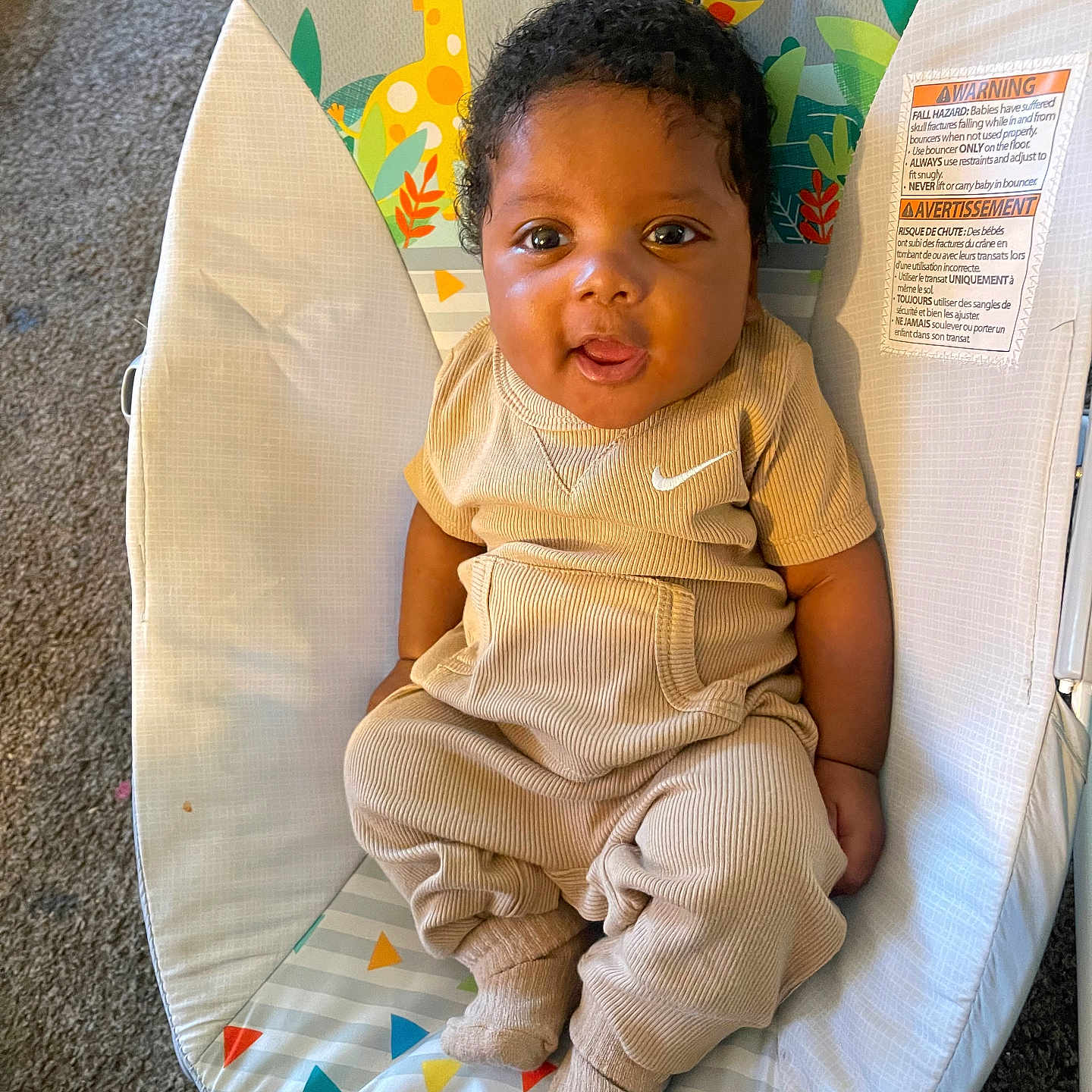 Demarion is registered to the contest to win money with this photo: baby, baby_seat, beige_clothing, carpet, child, colorful_background, comfort, curly_hair, cute, giraffe_pattern, happy, indoors, infant, playful, portrait, smiling, socks, soft_toys, tongue_out, young_child