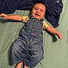 baby, child, denim_overalls, smiling, lying_down, indoor, bed_sheet, blanket, clothing, cute, happy, infant, person, face, skin, foot, hand, toddler, casual, portrait