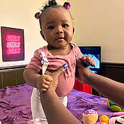Koemi is registered to the contest to win money with this photo: baby, child, girl, pink, hair_ties, hands, toys, blanket, bed, indoor, person, clothing, face, smile, wall, neon_sign, purple, white_pants, cute, holding
