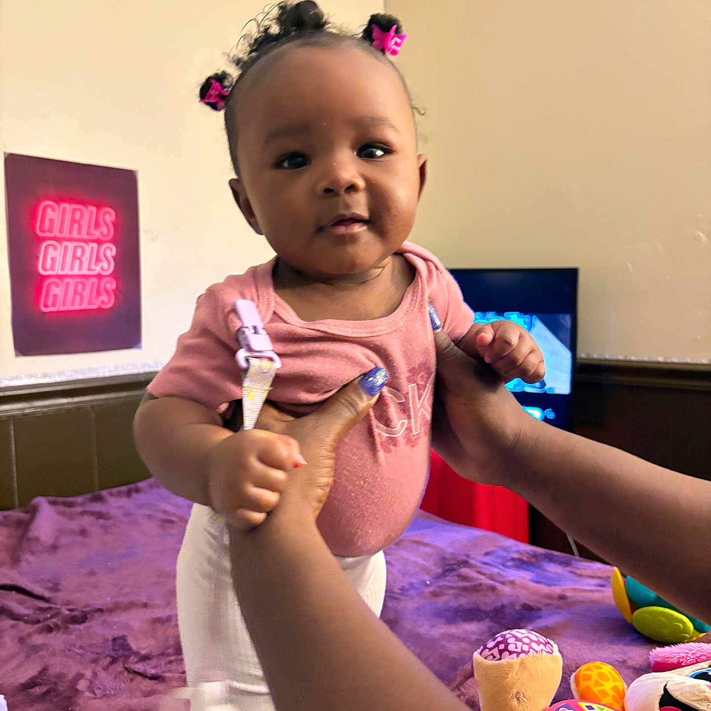 Koemi is registered to the contest to win money with this photo: baby, bed, blanket, child, clothing, cute, face, girl, hair_ties, hands, holding, indoor, neon_sign, person, pink, purple, smile, toys, wall, white_pants