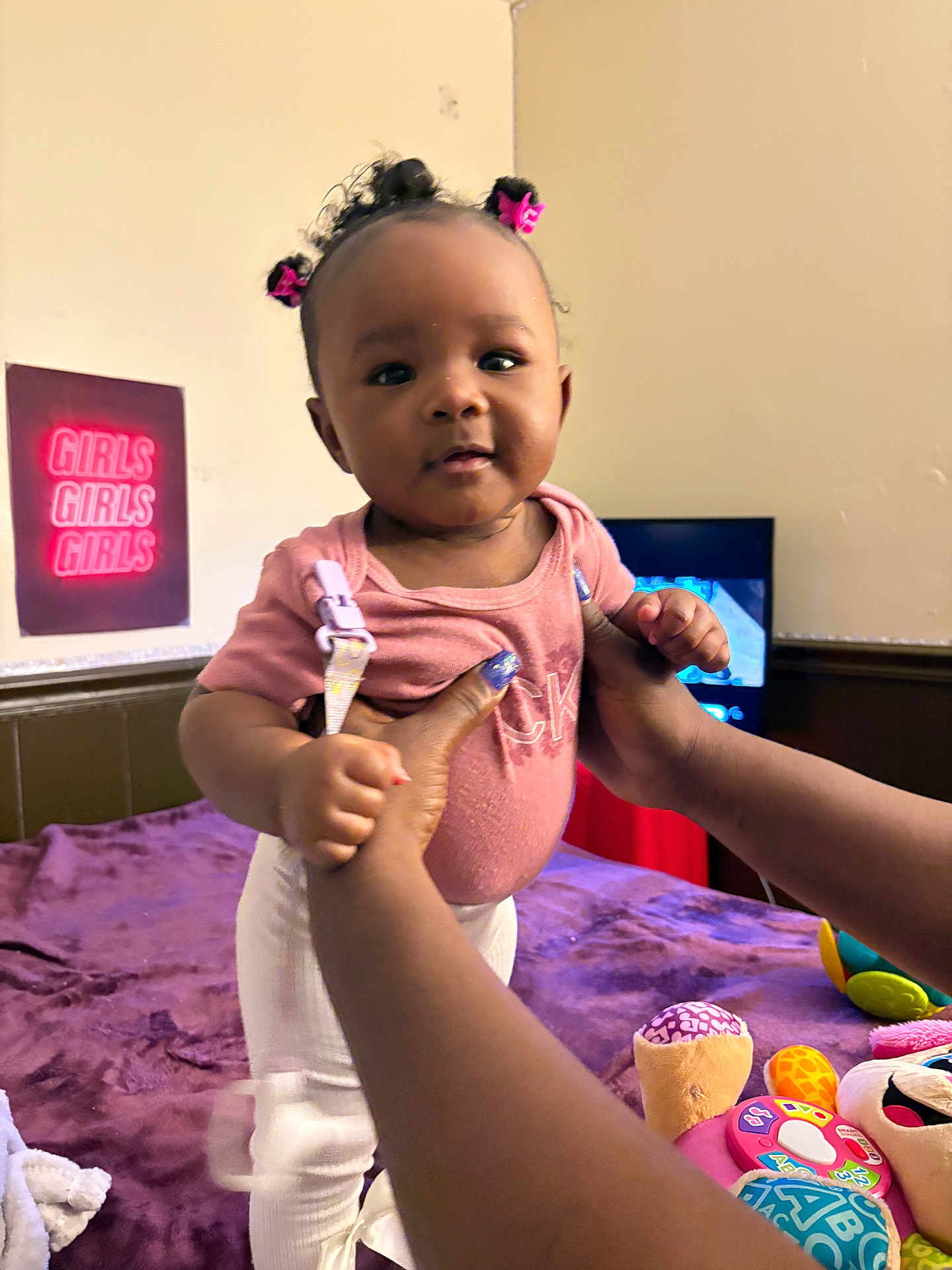 Koemi is registered to the contest to win money with this photo: baby, child, girl, pink, hair_ties, hands, toys, blanket, bed, indoor, person, clothing, face, smile, wall, neon_sign, purple, white_pants, cute, holding