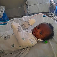 Josiah is registered to the contest to win money with this photo: baby, infant, hospital, crib, blanket, mittens, medical_tubes, face, person, bed, newborn, clothing, indoors, healthcare, patient, resting, cute, small, lying, hospital_room