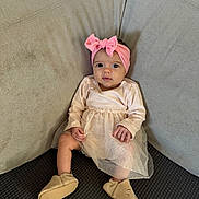 Lyvia a rejoint le concours — aidez-le/la à gagner de superbes lots ! baby, child, pink_headband, bow, dress, slippers, couch, seated, infant, cute, indoors, looking_at_camera, soft_fabric, cozy, small_hands, wide_eyes, light_skin, footwear, comfortable, home