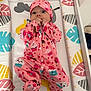 baby, infant, onesie, pink_clothing, cupcake_pattern, crib, mattress, colorful, leaf_pattern, animal_print, hand_to_mouth, cute, child, sleepwear, hat, footed_pajamas, portrait, indoors, young_child, playful