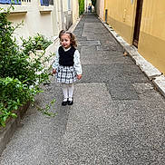 Lina participe au concours pour gagner de l'argent avec cette photo : alley, black_shoes, blue_windows, building, child, curly_hair, daytime, girl, greenery, long_sleeve_shirt, outdoor, pavement, plaid_skirt, plants, sidewalk, street, urban, walking, white_tights, yellow_wall
