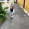 child, girl, street, alley, plants, greenery, yellow_wall, blue_windows, pavement, curly_hair, white_tights, black_shoes, plaid_skirt, long_sleeve_shirt, urban, outdoor, walking, daytime, building, sidewalk