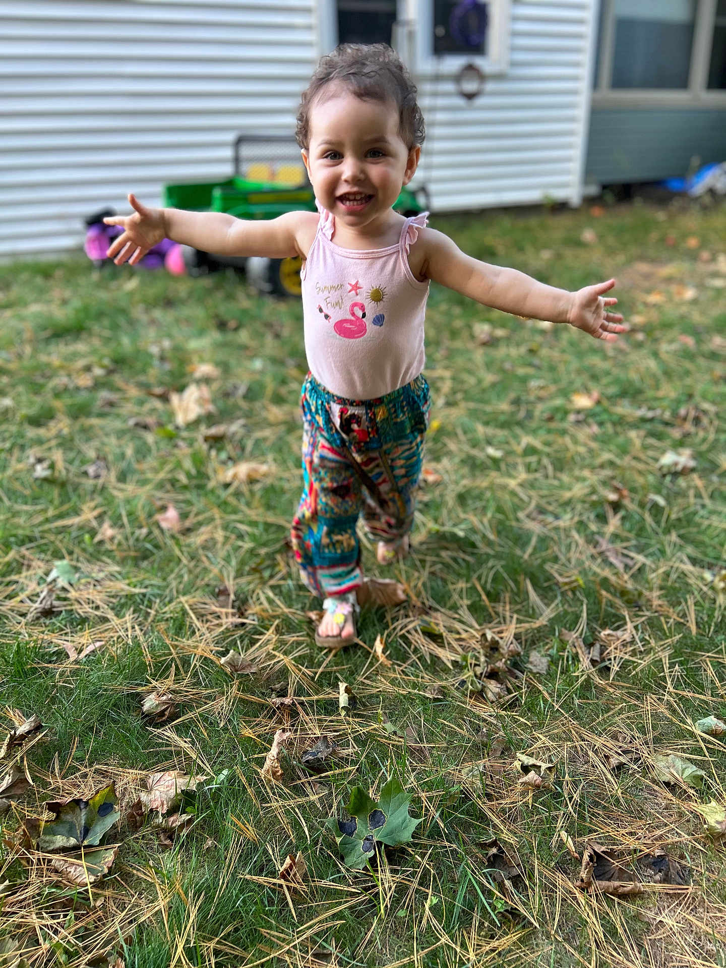 Rain joined the competition — help win amazing prizes! child, toddler, smiling, outdoor, grass, leaves, pine_needles, colorful_pants, pink_tank_top, happy, running, arms_outstretched, curly_hair, barefoot, summer_fun, yard, house, window, toy, daylight