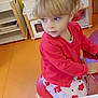 blonde_hair, blue_eyes, chair, child, childhood, cute, flower_dress, furniture, girl, indoor, kid, orange_floor, pigtails, playroom, portrait, red_cardigan, sitting, table, thoughtful, yellow_wall