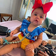 Kevrianys Sophia is registered to the contest to win money with this photo: baby, child, red_bow, colorful_clothing, table, hand, chair, room, indoor, person, watch, bracelet, television, picture_frame, curtain, pink_tablecloth, skin, face, expression, sitting
