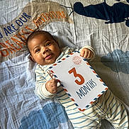 Freddie is registered to the contest to win money with this photo: baby, blanket, card, child, cloud, cute, face, hand, helicopter, indoors, infant, lying_down, milestone, pajamas, portrait, smile, soft_lighting, striped_clothing, text, young_child