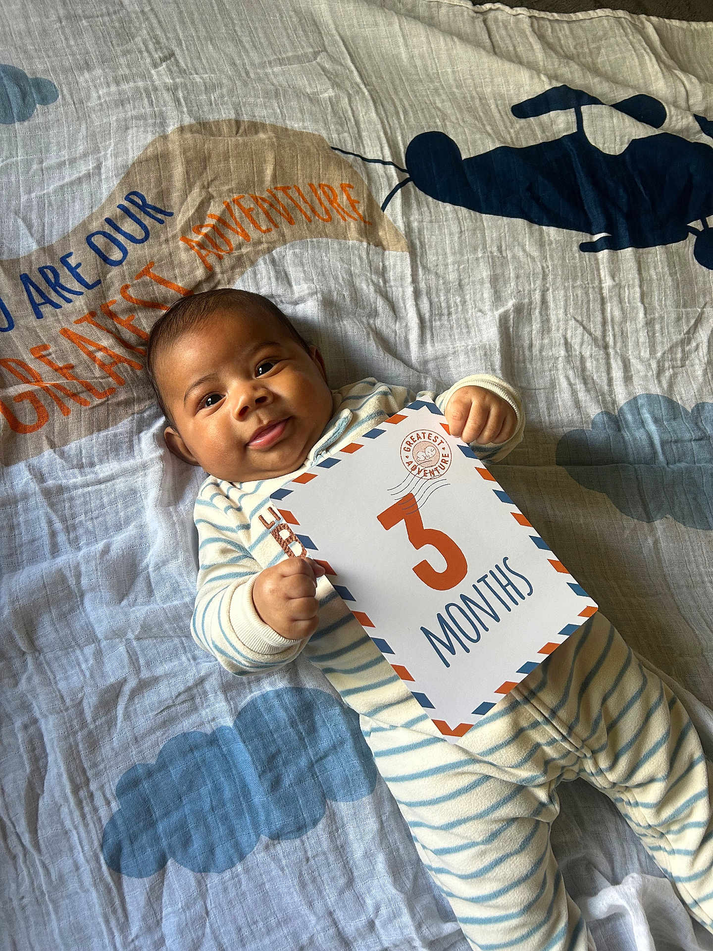 Freddie is registered to the contest to win money with this photo: baby, blanket, card, child, cloud, cute, face, hand, helicopter, indoors, infant, lying_down, milestone, pajamas, portrait, smile, soft_lighting, striped_clothing, text, young_child