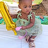 toddler, child, balloon, green_dress, table, toy, outdoor, party, birthday, decorations, cartoon_backdrop, sandals, curly_hair, cute, festive, celebration, young_child, sitting, holding, event