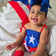 Emiely is registered to the contest to win money with this photo: baby, child, smiling, blue_bow, red_socks, puerto_rican_flag, flag, dress, happy, portrait, infant, cute, sitting, indoors, white_background, headwear, bright_colors, celebration, festive, person
