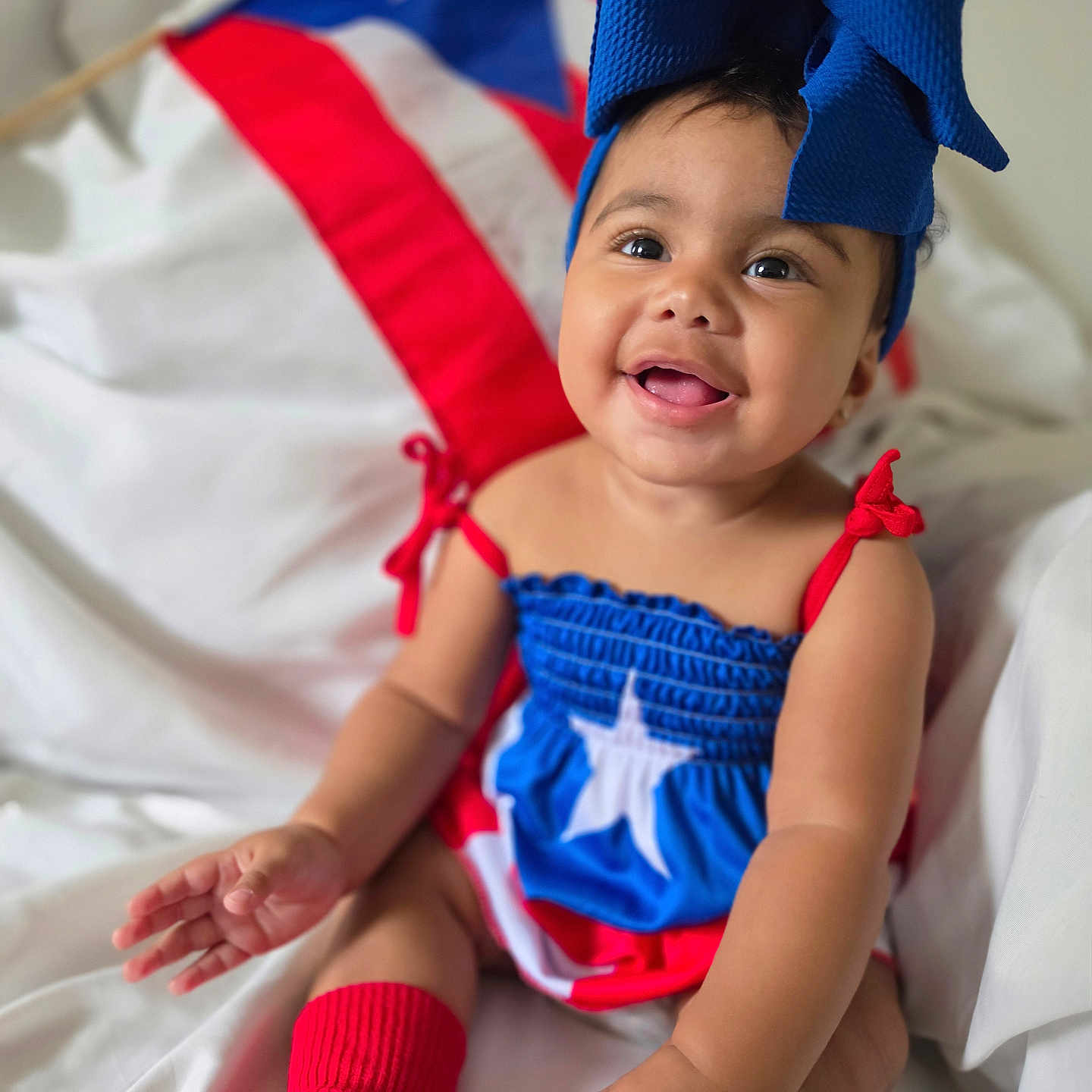 Emiely is registered to the contest to win money with this photo: baby, child, smiling, blue_bow, red_socks, puerto_rican_flag, flag, dress, happy, portrait, infant, cute, sitting, indoors, white_background, headwear, bright_colors, celebration, festive, person