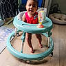 baby, walker, indoor, child, red_clothing, floor, wooden_floor, toy, happy, smiling, heart_graphics, curtains, dog_bed, dog, chair, person, feet, playful, cute, infant
