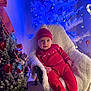 baby, child, red_clothing, hat, chair, fuzzy_blanket, christmas_tree, ornaments, lights, holiday, indoor, decorations, festive, seasonal, smiling, person, cozy, cute, toddler, winter