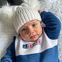 baby, child, hat, knit_hat, pom_poms, blue_sweatshirt, white_sweatshirt, jordan_logo, blanket, soft_texture, face, eyes, mouth, hand, furniture, cozy, indoor, portrait, cute, expression