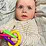 baby, infant, child, toy, plastic_rings, striped_clothing, bed, bedding, blanket, curious, face, hands, indoor, portrait, closeup, cute, young_child, soft_light, expression, lying_down