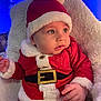 baby, santa_claus_costume, red_clothing, white_fur_trim, hat, infant, child, holiday, christmas, blue_lights, blanket, soft_texture, cute, portrait, indoors, festive, smiling, face, hand, cozy