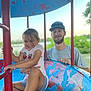 child, man, playground, playground_equipment, smiling, cap, sunglasses, shorts, tshirt, outdoor, daylight, holding, happy, grass, lake, trees, metal, rust, graffiti, people