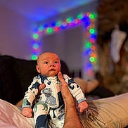 Jayce joined the competition — help win amazing prizes! baby, infant, onesie, animal_print, person, hand, adult, holding, couch, indoor, holiday_lights, blurred_background, cozy, festive, portrait, cute, wide_eyes, soft_lighting, relaxed, home