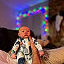 baby, infant, onesie, animal_print, person, hand, adult, holding, couch, indoor, holiday_lights, blurred_background, cozy, festive, portrait, cute, wide_eyes, soft_lighting, relaxed, home