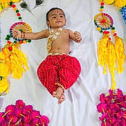 Yogi is registered to the contest to win money with this photo: baby, child, traditional_clothing, red_pants, gold_jewelry, flower_arrangement, yellow_flowers, pink_flowers, white_sheet, decorative_wheels, peacock_feathers, smiling, lying_down, indoor, portrait, cute, celebration, cultural, festive, infant