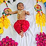 baby, child, traditional_clothing, red_pants, gold_jewelry, flower_arrangement, yellow_flowers, pink_flowers, white_sheet, decorative_wheels, peacock_feathers, smiling, lying_down, indoor, portrait, cute, celebration, cultural, festive, infant