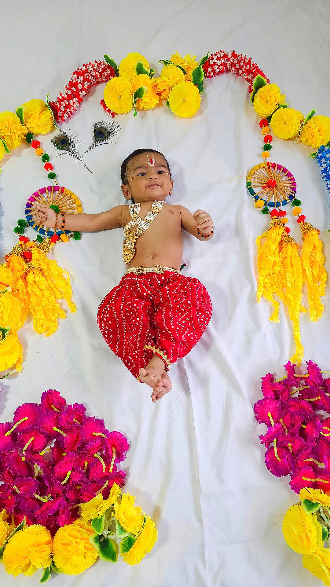 Yogi is registered to the contest to win money with this photo: baby, child, traditional_clothing, red_pants, gold_jewelry, flower_arrangement, yellow_flowers, pink_flowers, white_sheet, decorative_wheels, peacock_feathers, smiling, lying_down, indoor, portrait, cute, celebration, cultural, festive, infant