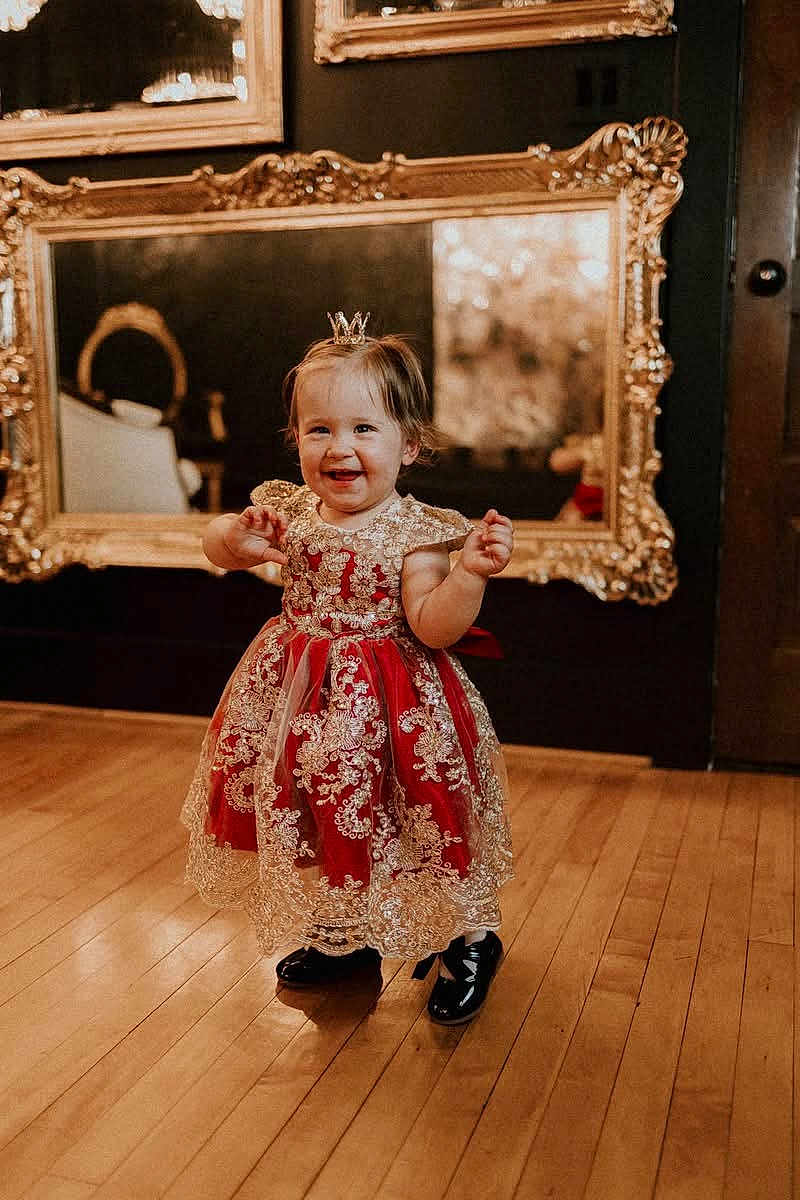Svetlana is registered to the contest to win money with this photo: toddler, child, dress, red_dress, gold_lace, crown, smiling, standing, wooden_floor, mirror, gold_frame, indoor, happy, person, footwear, black_shoes, cute, portrait, decor, elegant