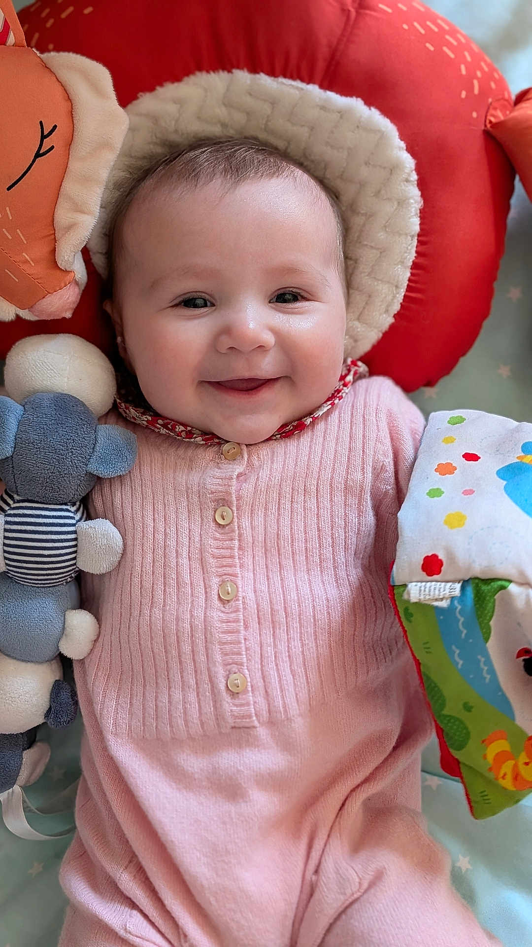Victoire participe au concours pour gagner de l'argent avec cette photo : baby, infant, smiling, face, cheeks, eyes, onesie, pink_clothing, plush_toy, stuffed_animal, pillow, headrest, blanket, toy_book, buttons, cozy, closeup, portrait, nursery, happy