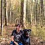 person, dog, forest, trail, trees, outdoor, nature, leash, harness, crouching, smiling, black_dog, casual_clothing, shorts, tshirt, glasses, daylight, leaves, path, friendship
