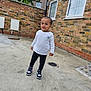 toddler, child, outdoor, concrete, brick_wall, window, tree, sneakers, white_shirt, black_pants, standing, curious, casual_clothing, young_child, person, daylight, sidewalk, urban, young_boy, smile