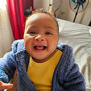 Adriano is registered to the contest to win money with this photo: baby, child, smiling, blue_jacket, yellow_shirt, indoor, bed, fan, curtains, person, happy, portrait, fuzzy_clothing, infant, cute, warm, cozy, young_child, human, closeup