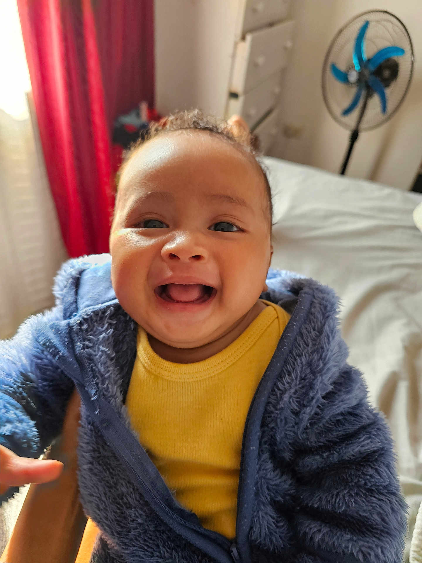Adriano is registered to the contest to win money with this photo: baby, child, smiling, blue_jacket, yellow_shirt, indoor, bed, fan, curtains, person, happy, portrait, fuzzy_clothing, infant, cute, warm, cozy, young_child, human, closeup