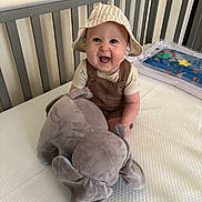 Mikey joined the competition — help win amazing prizes! baby, child, smiling, hat, bucket_hat, plush_toy, elephant, crib, nursery, overalls, white_wall, mattress, water_play_mat, happy, sitting, indoor, cute, person, infant, toy