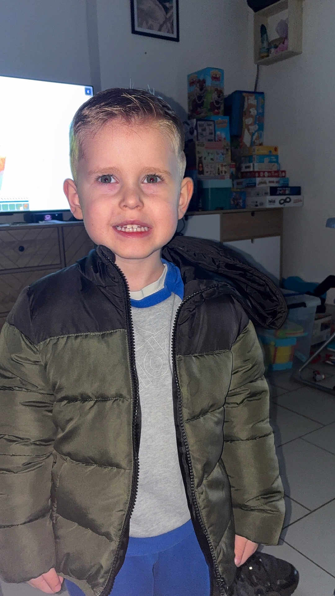 Benji participe au concours pour gagner de l'argent avec cette photo : child, boy, young_boy, smiling, teeth, jacket, puffer_jacket, indoor, living_room, toys, tv_screen, tile_floor, dresser, hair, ears, eyes, pants, flash_photography, portrait, casual_clothing