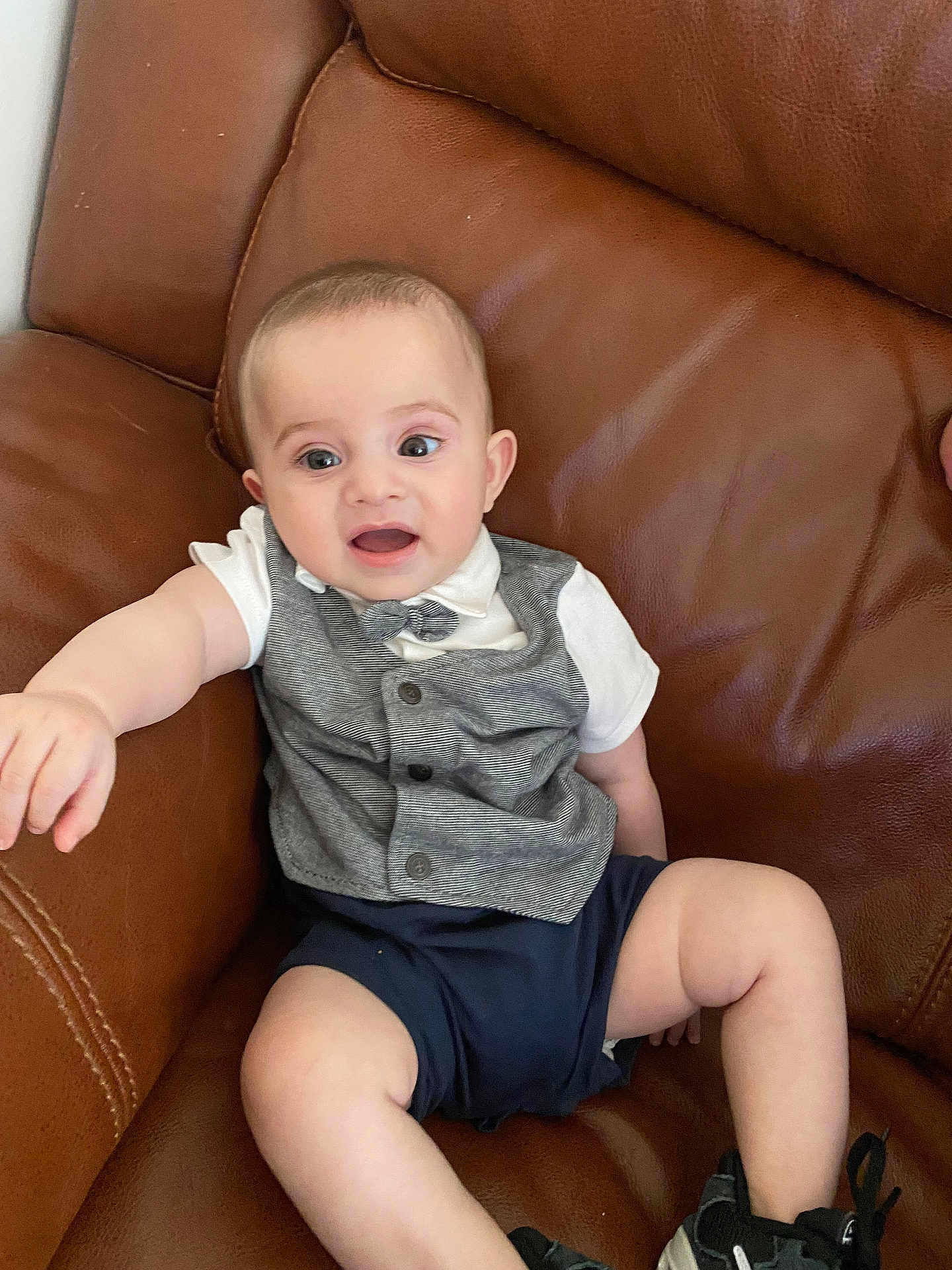Muhammed joined the competition — help win amazing prizes! baby, infant, child, couch, leather, clothing, vest, bow_tie, shorts, sneakers, person, sitting, furniture, indoor, expression, reaching, cute, small, happy, face