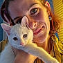 affection, animal, cat, closeup, cute, earring, face, feline, hand, indoor, kitten, person, pet, piercing, portrait, smile, striped_shirt, warm_lighting, white_cat, yellow_shirt
