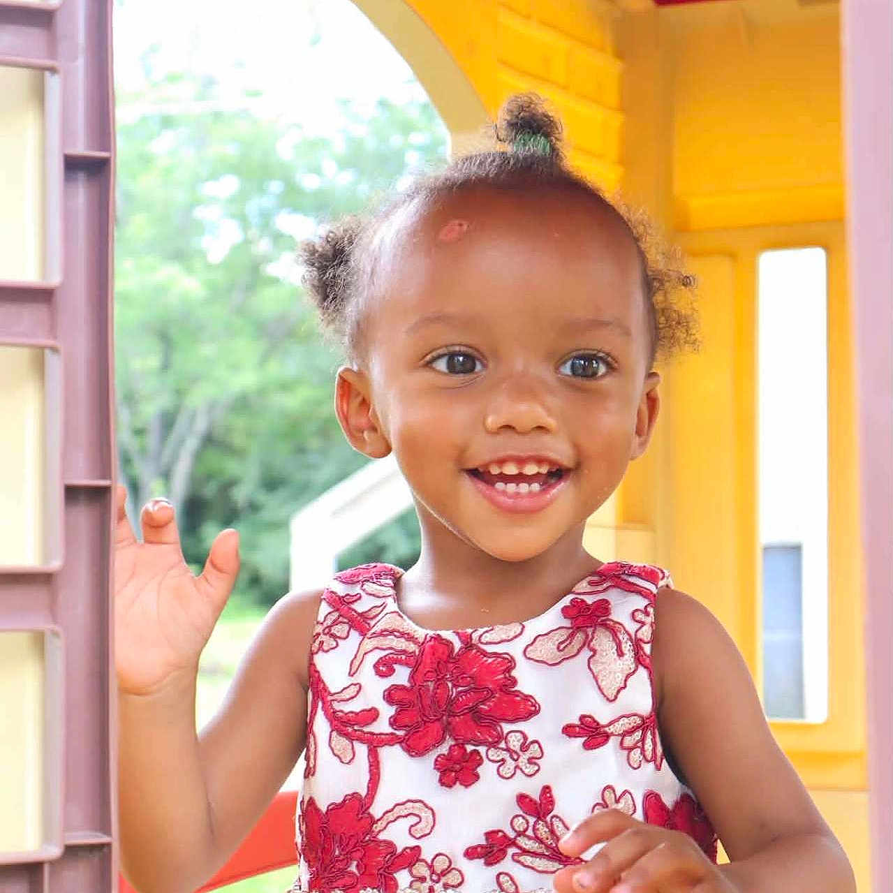 An'Dreya is registered to the contest to win money with this photo: child, girl, toddler, smiling, waving, portrait, face, eyes, teeth, hair_bun, curly_hair, floral_dress, dress, playhouse, window, colorful, yellow, outdoor, daylight, happy