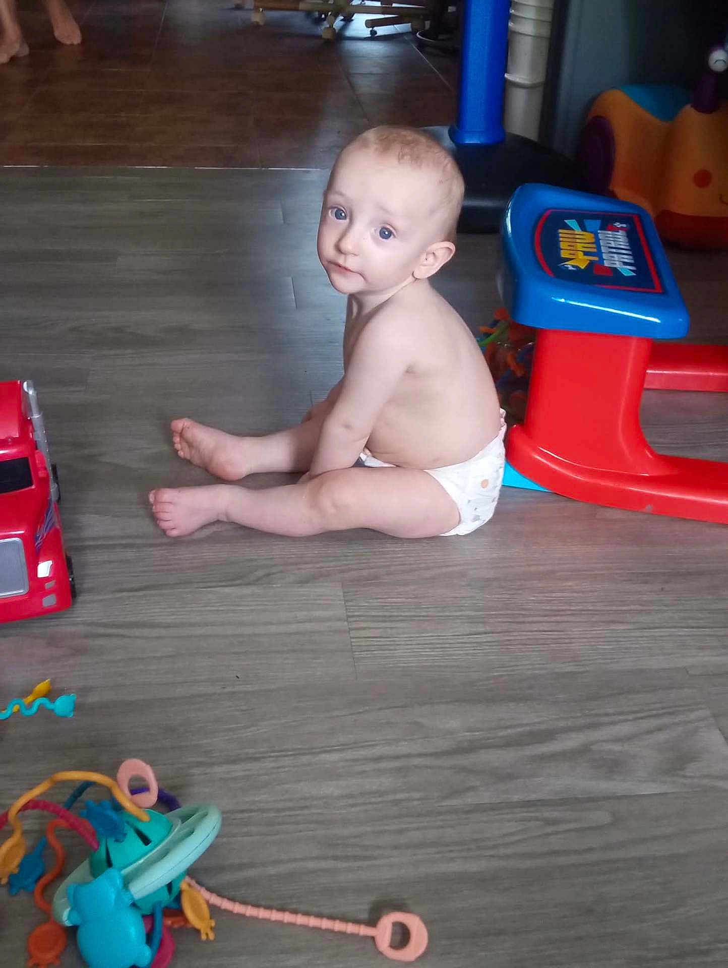 Braxtan is registered to the contest to win money with this photo: toddler, child, diaper, floor, toy, plastic, play_table, red, blue, curious, indoor, wooden_floor, person, baby, sitting, looking_back, playtime, colorful, home, young_child