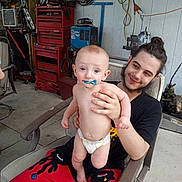 Braxtan joined the competition — help win amazing prizes! baby, diaper, pacifier, man, smile, chair, workshop, tools, garage, casual_clothing, red_pants, hair_tied, beard, holding, curious, indoor, concrete_floor, equipment, young_adult, child