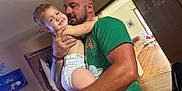 Bryson is registered to the contest to win money with this photo: arm, bracelet, child, couch, diaper, door, facial_hair, family, green_tshirt, holding, home, indoor, light_fixture, man, shelf, smile, tattoo, toddler, window, wooden_floor