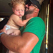 Bryson is registered to the contest to win money with this photo: affection, arm, beard, cap, casual_clothing, child, closeup, curtains, diaper, face, family, green_shirt, holding, indoor, man, natural_light, skin, sleepy, toddler, window