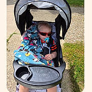 Marcus is registered to the contest to win money with this photo: child, baby, toddler, stroller, sunglasses, red_sunglasses, pacifier, blanket, spiderman, outdoor, sunlight, sidewalk, grass, pavement, portrait, sitting, vehicle, infant, family_photo, screenshot