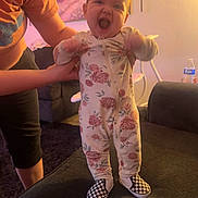 Lakelynn joined the competition — help win amazing prizes! baby, child, person, standing, onesie, floral_pattern, shoes, checkered, couch, living_room, adult_hand, smiling, happy, indoors, carpet, painting, water_bottle, furniture, home, warm_lighting