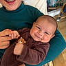 baby, child, smile, braces, holding, teddy_bear_patch, brown_outfit, teal_top, hand_holding, indoor, happy, face, person, furniture, chair, floor, wooden_floor, couch, plush_toy, diapers