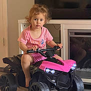 Noor is registered to the contest to win money with this photo: child, toddler, girl, portrait, indoor, living_room, pink_clothing, pink_atv, ride_on_toy, toy_vehicle, quad_bike, handlebars, wheels, fireplace, television, wood_floor, seated, curly_hair, serious_expression, family_home