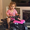 child, toddler, girl, portrait, indoor, living_room, pink_clothing, pink_atv, ride_on_toy, toy_vehicle, quad_bike, handlebars, wheels, fireplace, television, wood_floor, seated, curly_hair, serious_expression, family_home