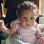 baby, child, curly_hair, blue_eyes, pink_shirt, striped_pants, blanket, indoor, soft_light, wooden_wall, shelf, framed_pictures, figurines, person, cute, infant, cozy, portrait, seated, curious
